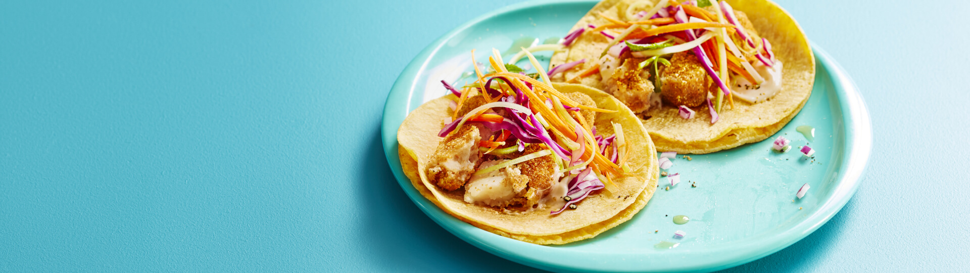 Aqua blue background with white plate topped with two fish tacos with cabbage and carrot coleslaw.