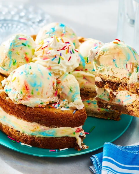 a hand holding an ice cream scoop over a tub of Compliments birthday cake flavour ice cream, pulling it through to scoop out white and coloured sprinkle scoop of ice cream, with cones ona plate in the background