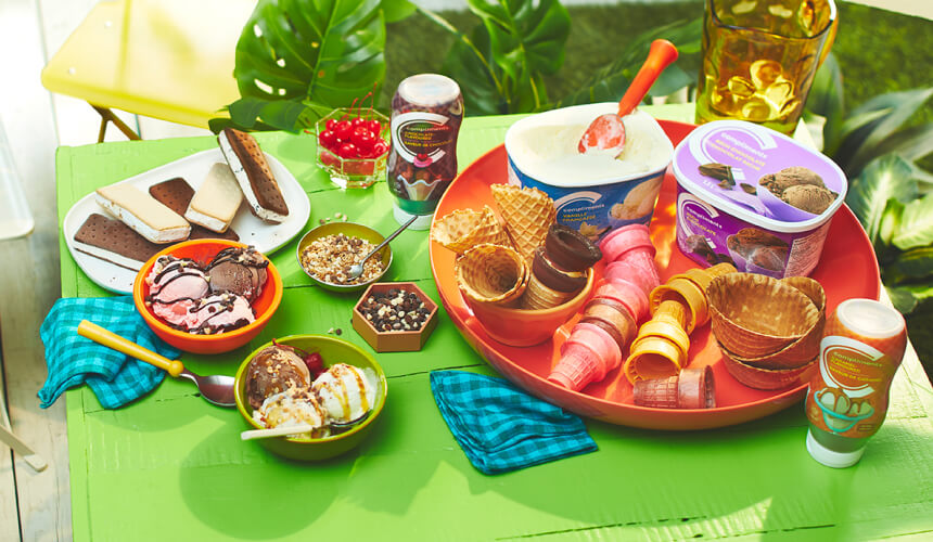 Green outdoor table with an array of ice cream sundae ingredients including ice cream, sundae sauces, cones, and toppings