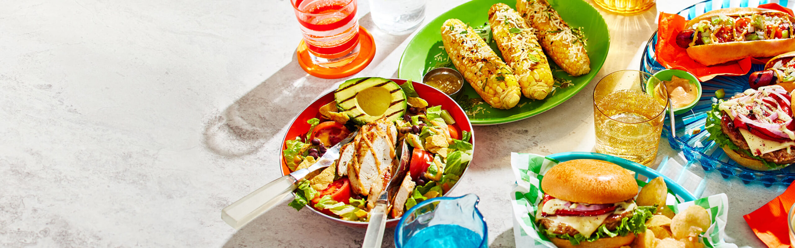 Grilled corn, grilled salad, burgers, and hot dogs on a white backdrop